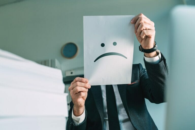 Unhappy businessman holding paper with frowning emoticon in office interior